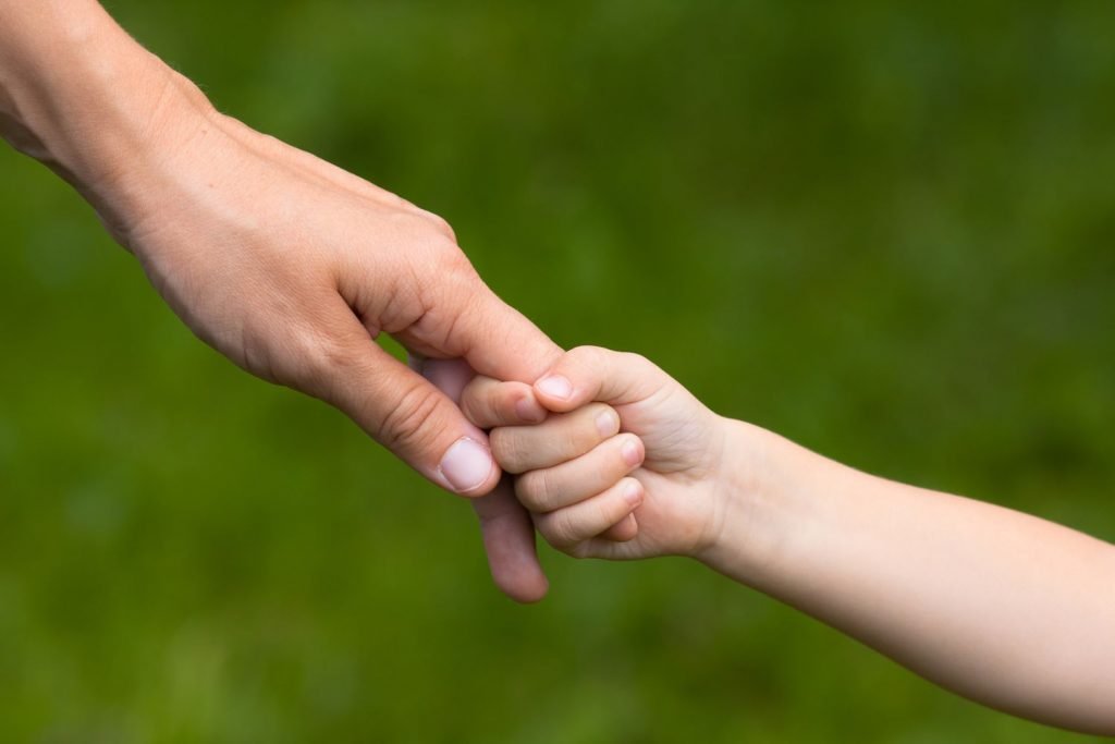 An adult holding a child's hand against a leafy backdrop.