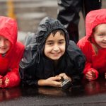 Three giggling children lying on their soaking wet school grounds in full wet weather gear.