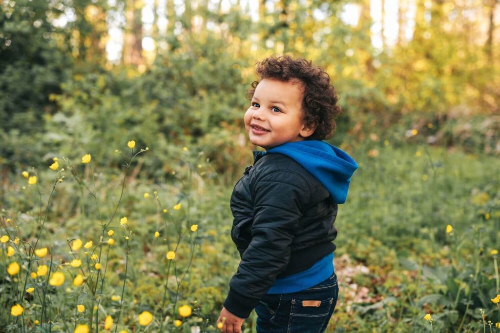 A toddler dressed for the outdoors laughing in sunny, green woodland.