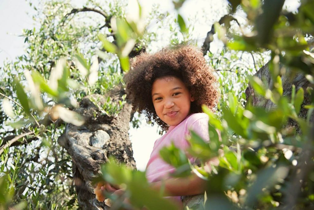 A young black girl smiling at the camera from her perch in a leafy tree she has climbed.