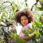 A young black girl smiling at the camera from her perch in a leafy tree she has climbed.