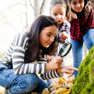 Three adolescent girls examining a tree trunk with a magnifying glass during an outdoor lesson.