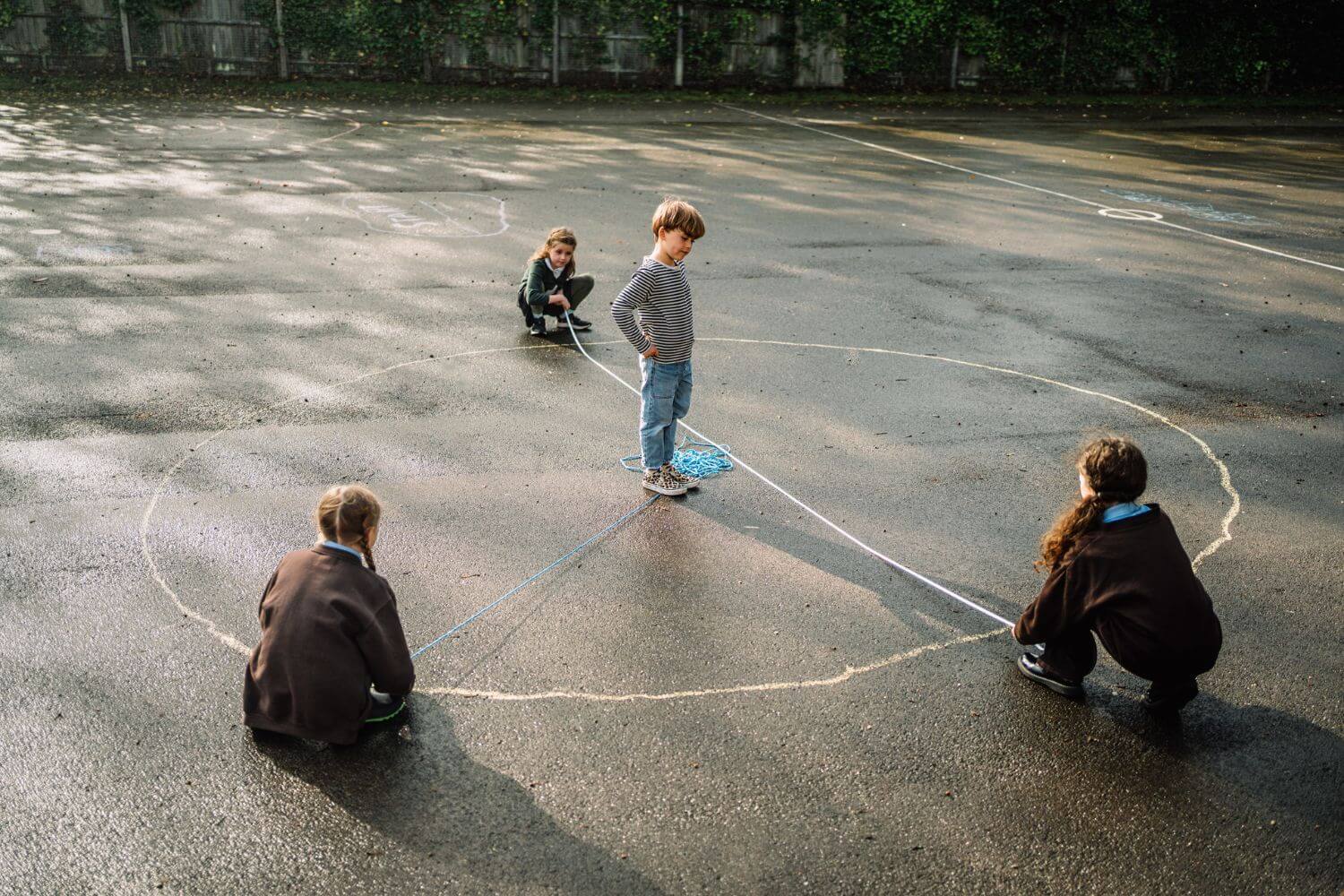 Primary school children using rope and chalk to draw a circle on the playground during an outdoor numeracy activity.