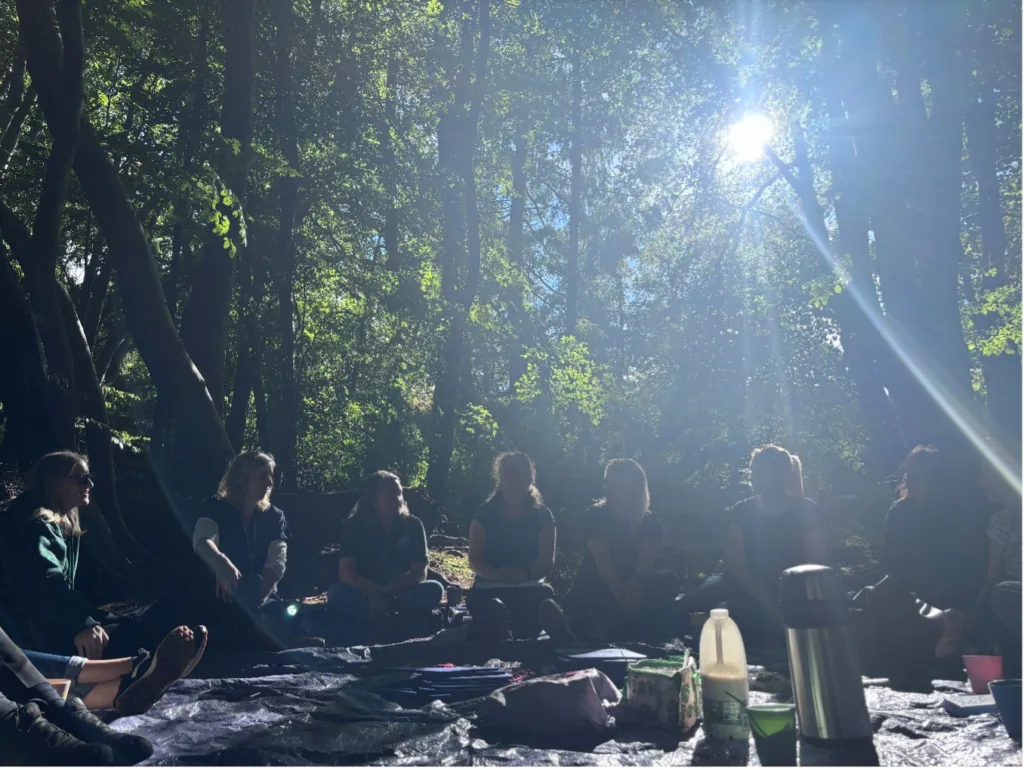 Adults sitting on tarpaulins in the woods with tea-making equipment in the middle