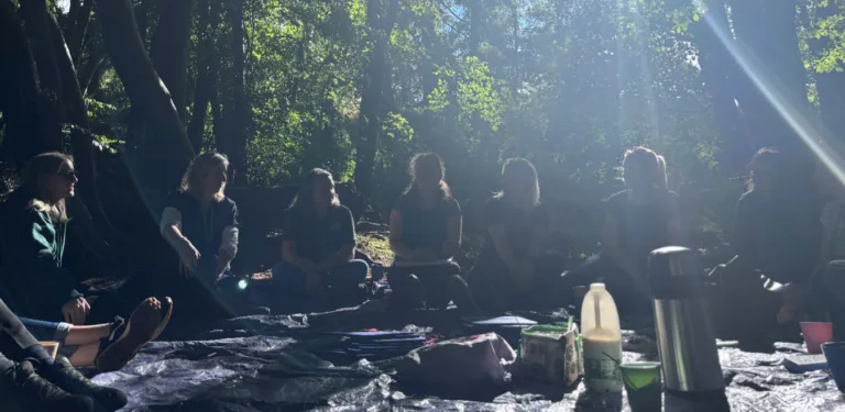 Adults sitting on tarpaulins in the woods with tea-making equipment in the middle